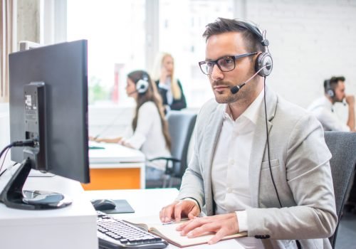 Serious man wearing formal clothes and headset looking at computer screen in bright office